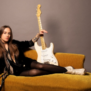 portrair de Nina Attal allongée sur un canapé brun avec sa guitare électrique blanche à la main