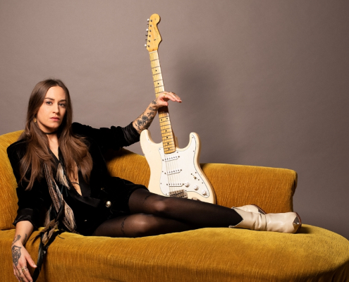portrair de Nina Attal allongée sur un canapé brun avec sa guitare électrique blanche à la main