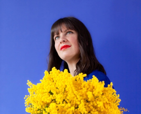 portrait sur fond bleu de l'artiste June Mio tenant un gros bouquet jaune